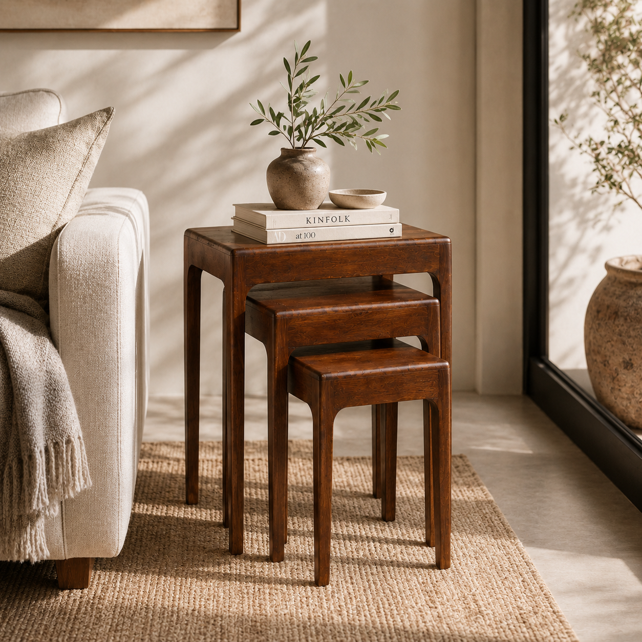 Three nested wooden nesting side tables in a warm medium brown finish, styled in a cozy living room next to a cream sofa with a vase and books on top.